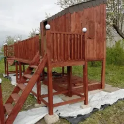 Drewniany plac zabaw dla dzieci z domkiem na podwyższeniu, schodami i balustradą, pomalowany na brązowo, z elementami oświetleniowymi w postaci kulistych lamp, otoczony zielenią i zabezpieczony folią.