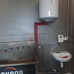 Montaż bojlera TESY nad umywalką z widocznymi instalacjami hydraulicznymi i narzędziami, w pomieszczeniu o ścianach z blachy trapezowej.