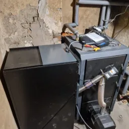 Nowoczesny piec CO z podajnikiem, z widocznymi rurami instalacji grzewczej, umieszczony w pomieszczeniu z surowymi ścianami.