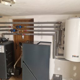 Hydr-All Lucjan Kruczyński - Kotłownia z piecem na paliwo stałe, zasobnikiem ciepłej wody Ferroli i izolowanymi rurami. Widoczne pompy obiegowe i zawory. Ujęcie w pomieszczeniu z drewnianymi drzwiami.