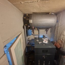 Hydr-All Lucjan Kruczyński - Szary piec gazowy Ferroli z podłączonym zasobnikiem wody w kotłowni. Widoczne rury, manometr i osoba podczas instalacji.