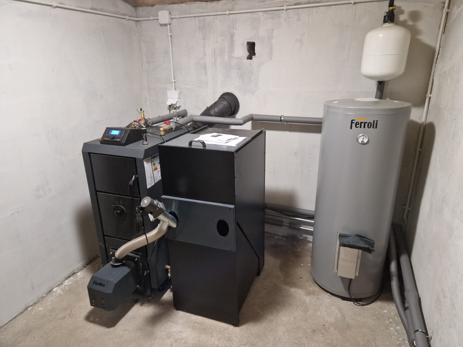 Kotłownia z piecem na paliwo stałe (ekogroszek) i zasobnikiem ciepłej wody użytkowej marki Ferolli. Widoczne rury instalacji CO i CWU.