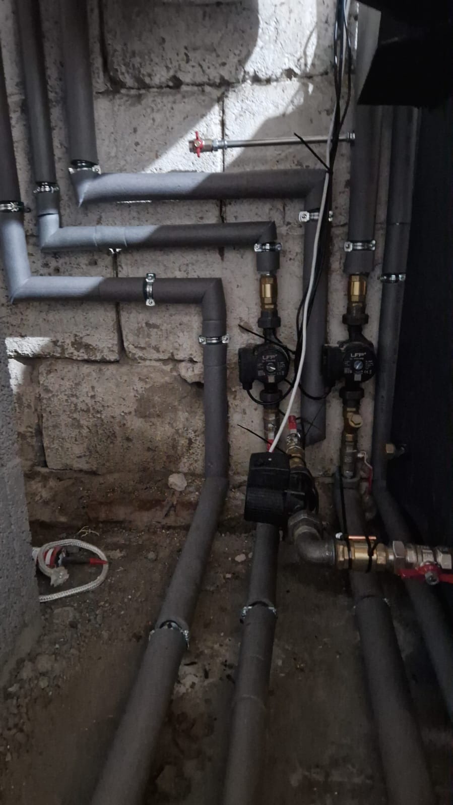 Szare rury instalacji CO z izolacją i pompami obiegowymi na tle starej, betonowej ściany. Widoczne zawory i elementy mocujące.