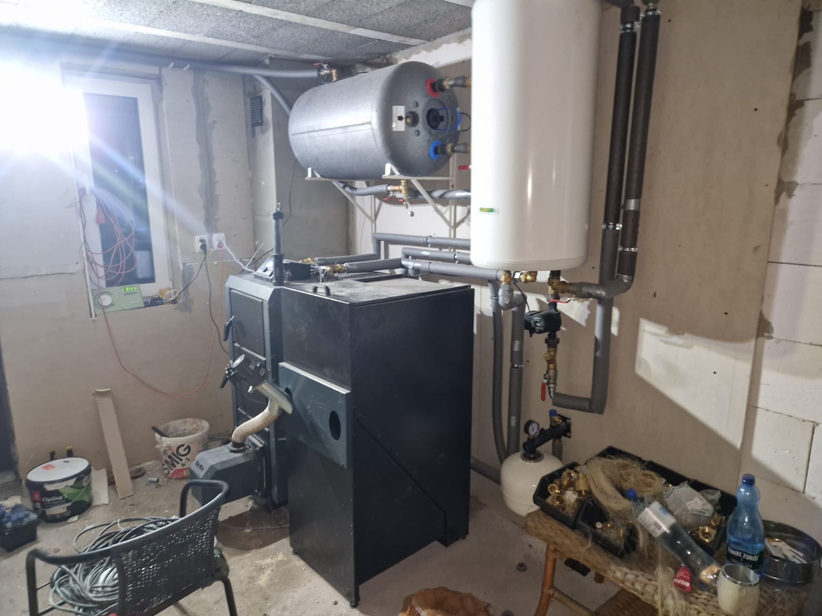 Kotłownia z piecem na paliwo stałe, zbiornikiem akumulacyjnym i bojlerem. Szare rury instalacji grzewczej. Surowe ściany z betonu komórkowego w tle.