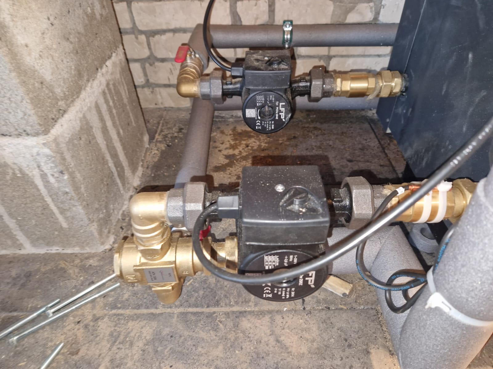 Dwie czarne pompy obiegowe LFP zamontowane na rurach z mosiężnymi złączkami, otulone szarą izolacją, na tle surowej ściany z cegły i betonowej podłogi.