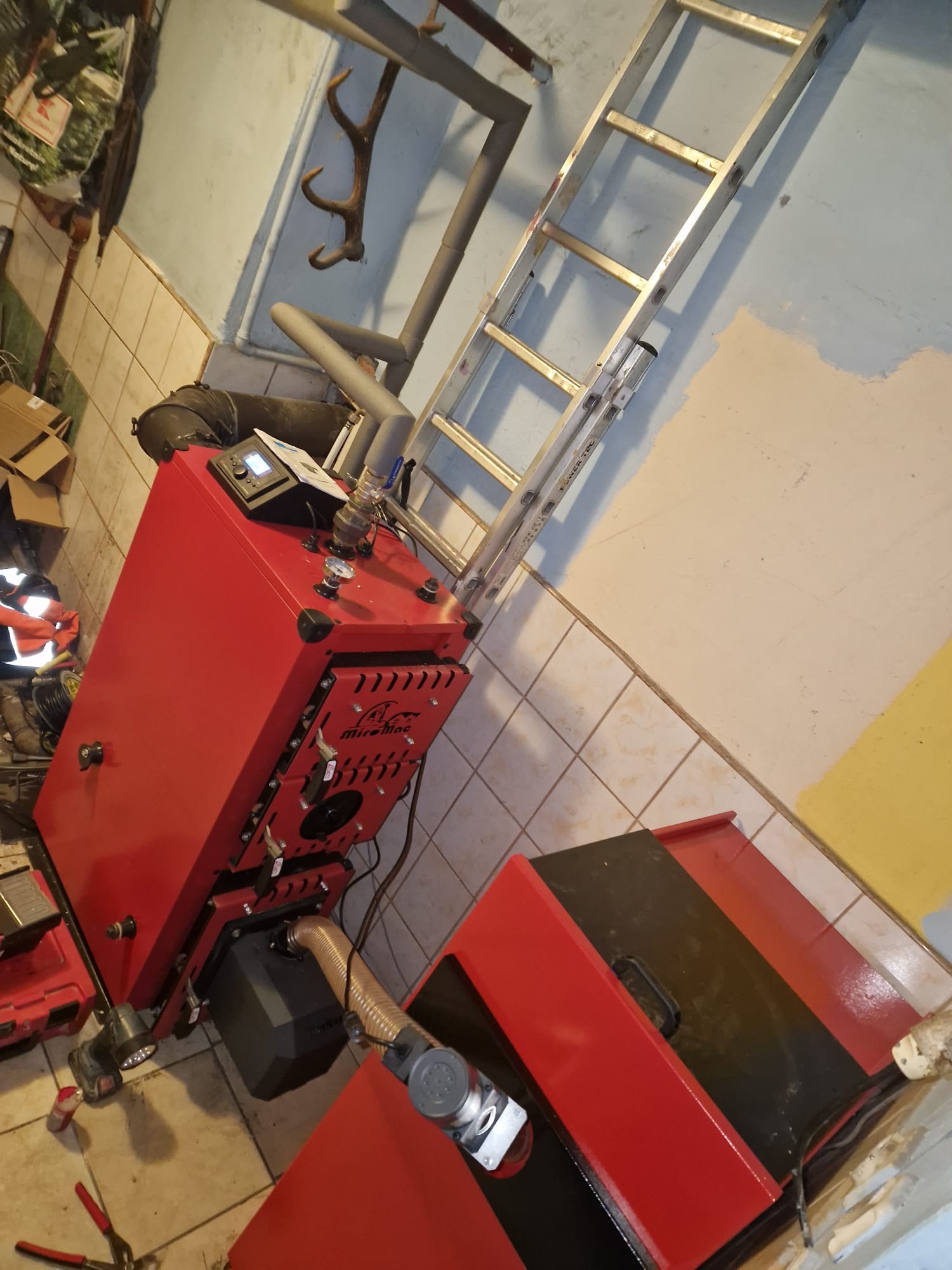 Czerwony piec CO marki MirellMac w kotłowni, z drabiną opartą o ścianę. Widoczne rury instalacji grzewczej i narzędzia hydrauliczne. Na ścianie wieszak na poroże.
