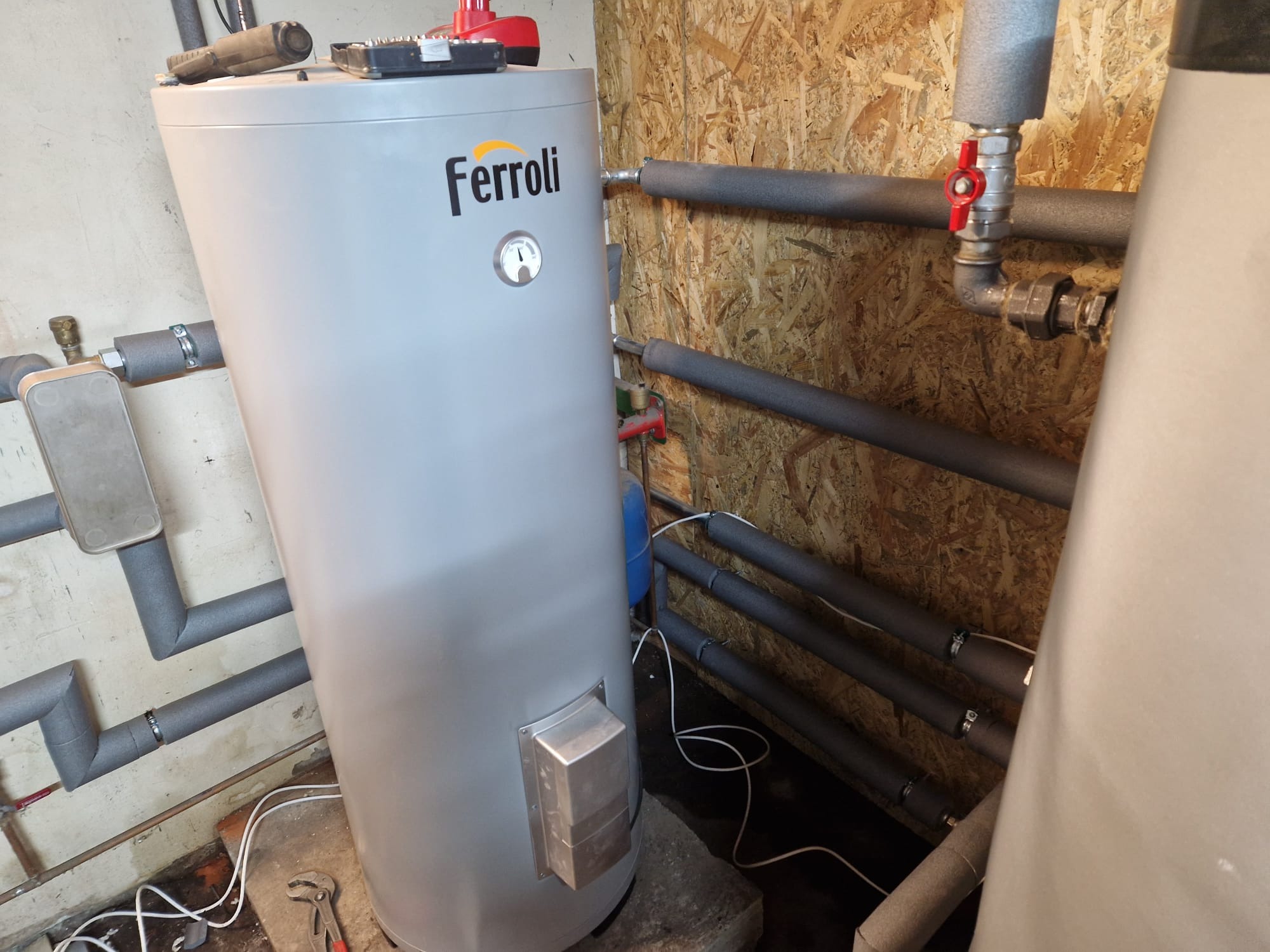 Zamontowany piec Ferroli w Koszalinie. Widoczne rury z izolacją termiczną i narzędzia. Piec stoi na betonowych bloczkach.