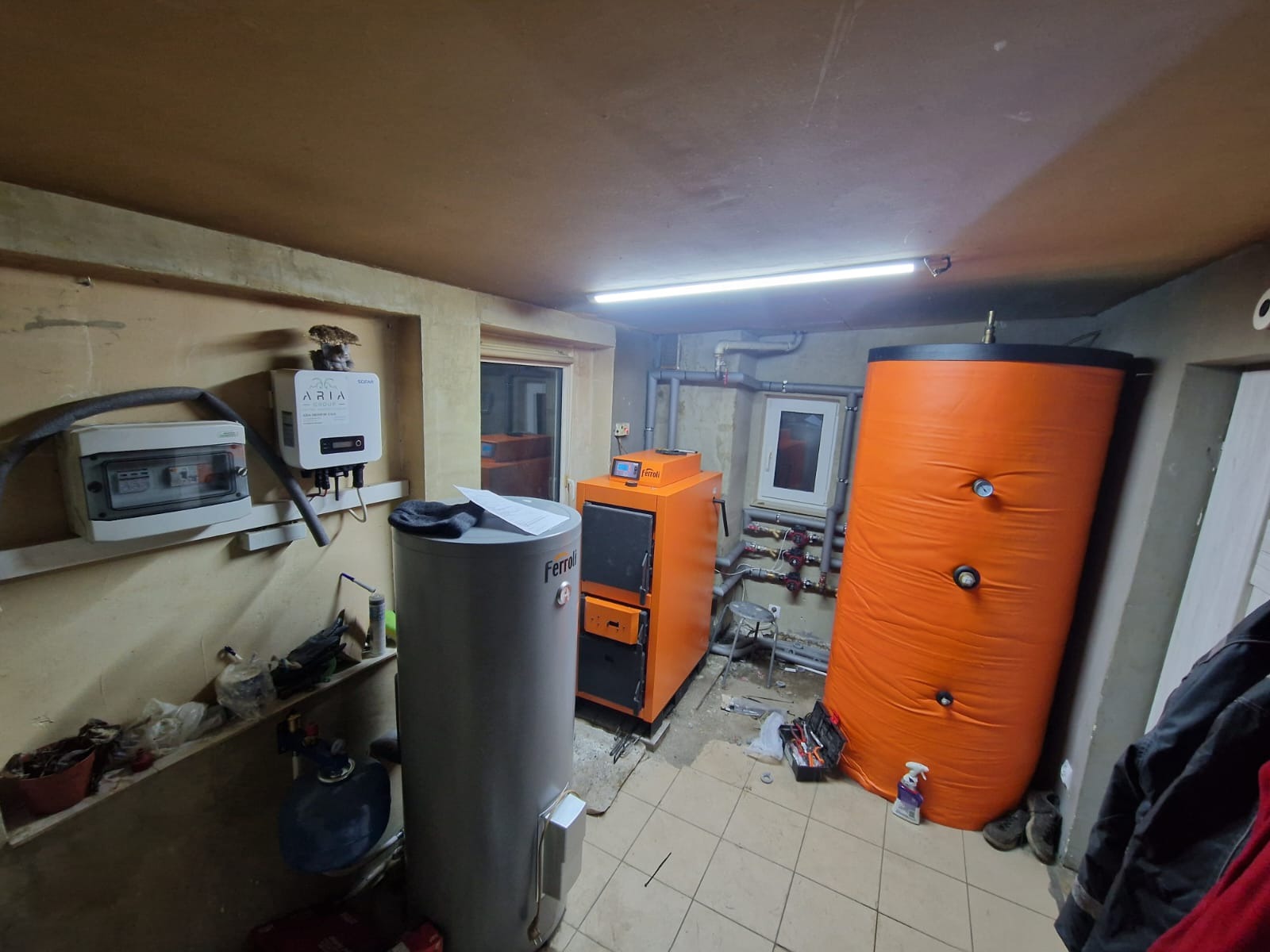 Pomieszczenie kotłowni z piecem Ferroli, zbiornikiem akumulacyjnym i instalacją CO. Widoczne rury, armatura i osprzęt. Nierówna ściana i podłoga z płytek.