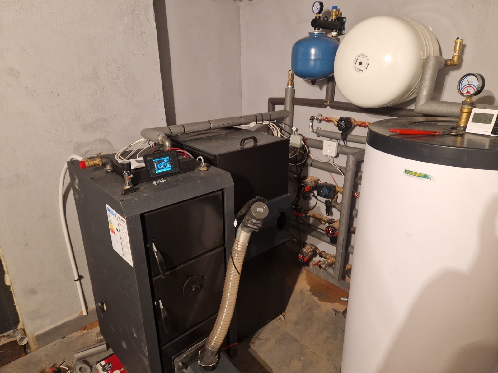 Kotłownia z piecem na paliwo stałe, zbiornikiem akumulacyjnym i instalacją CO. Widoczne rury, zawory i sterownik. Szare ściany, surowy stan wykończenia.