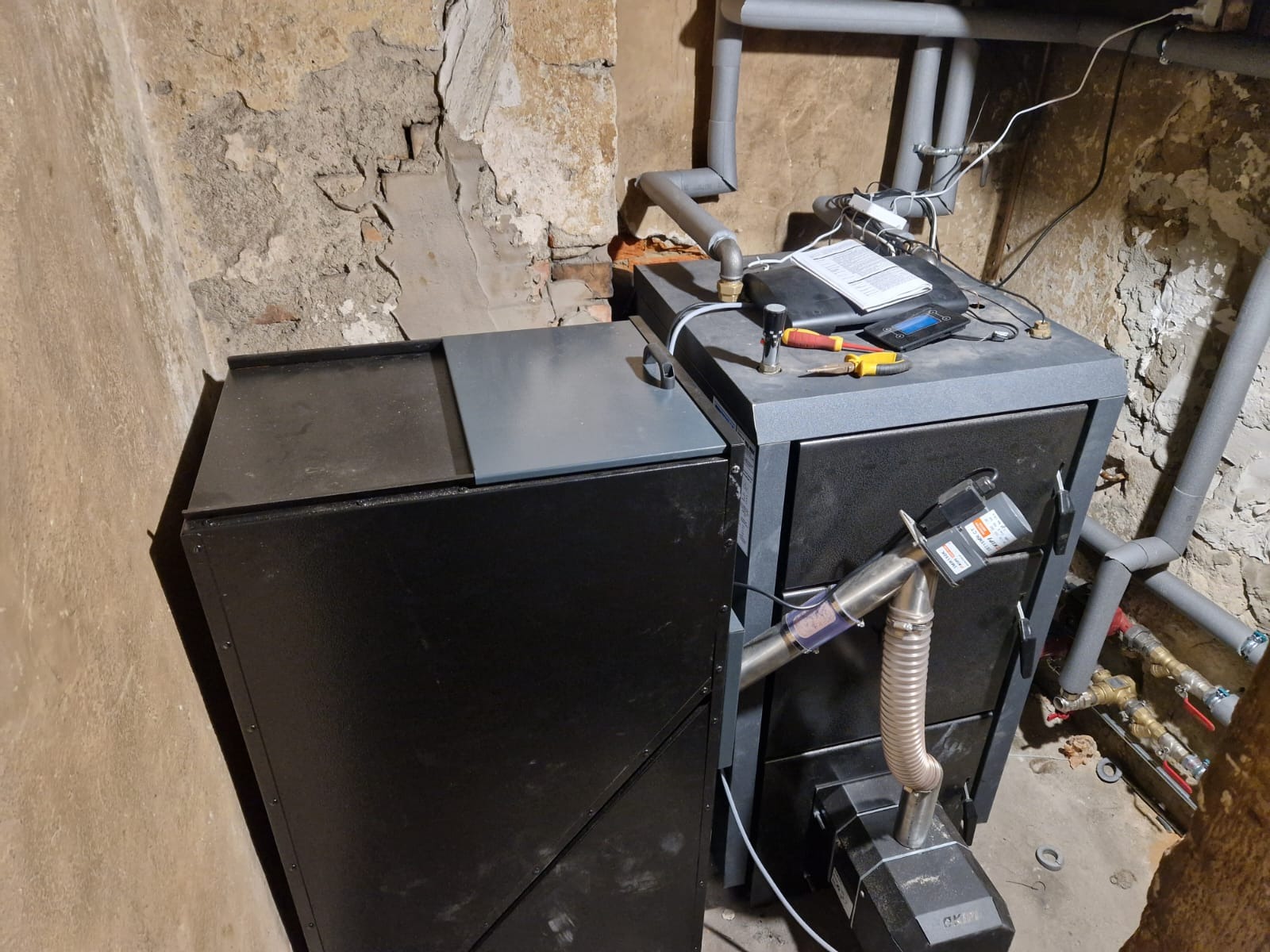 Nowoczesny piec CO z podajnikiem, z widocznymi rurami instalacji grzewczej, umieszczony w pomieszczeniu z surowymi ścianami.