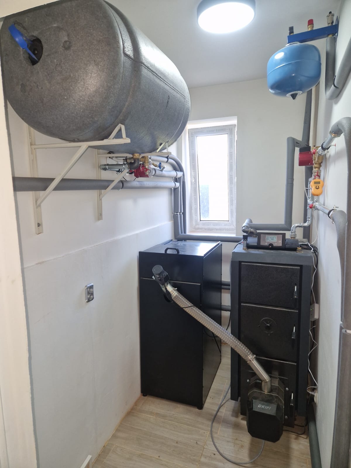 Zmontowana instalacja grzewcza w kotłowni: zbiornik akumulacyjny, piec na paliwo stałe z podajnikiem i osprzęt. Widoczne rury, zawory i sterownik.