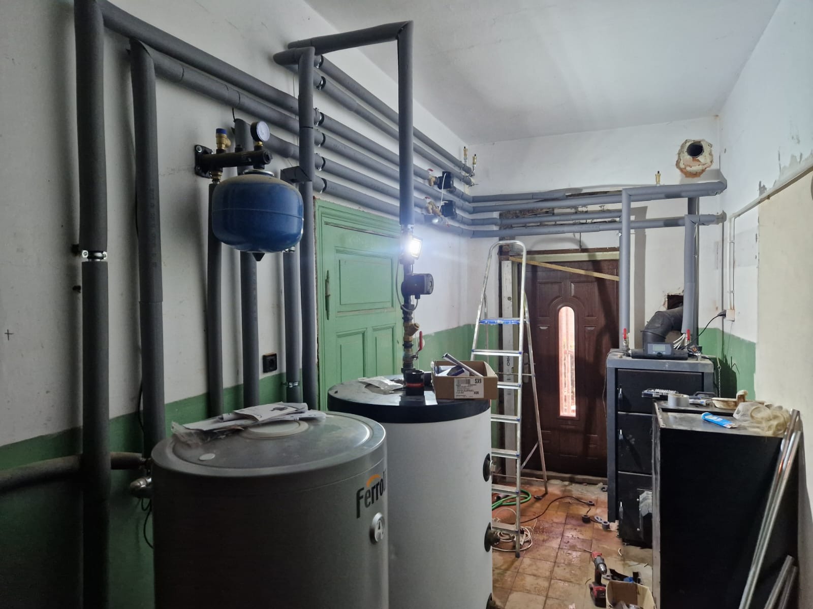 Wnętrze kotłowni w trakcie modernizacji: rury, zbiorniki Ferroli, drabina. Widoczne narzędzia i elementy instalacji grzewczej. Oświetlenie robocze.