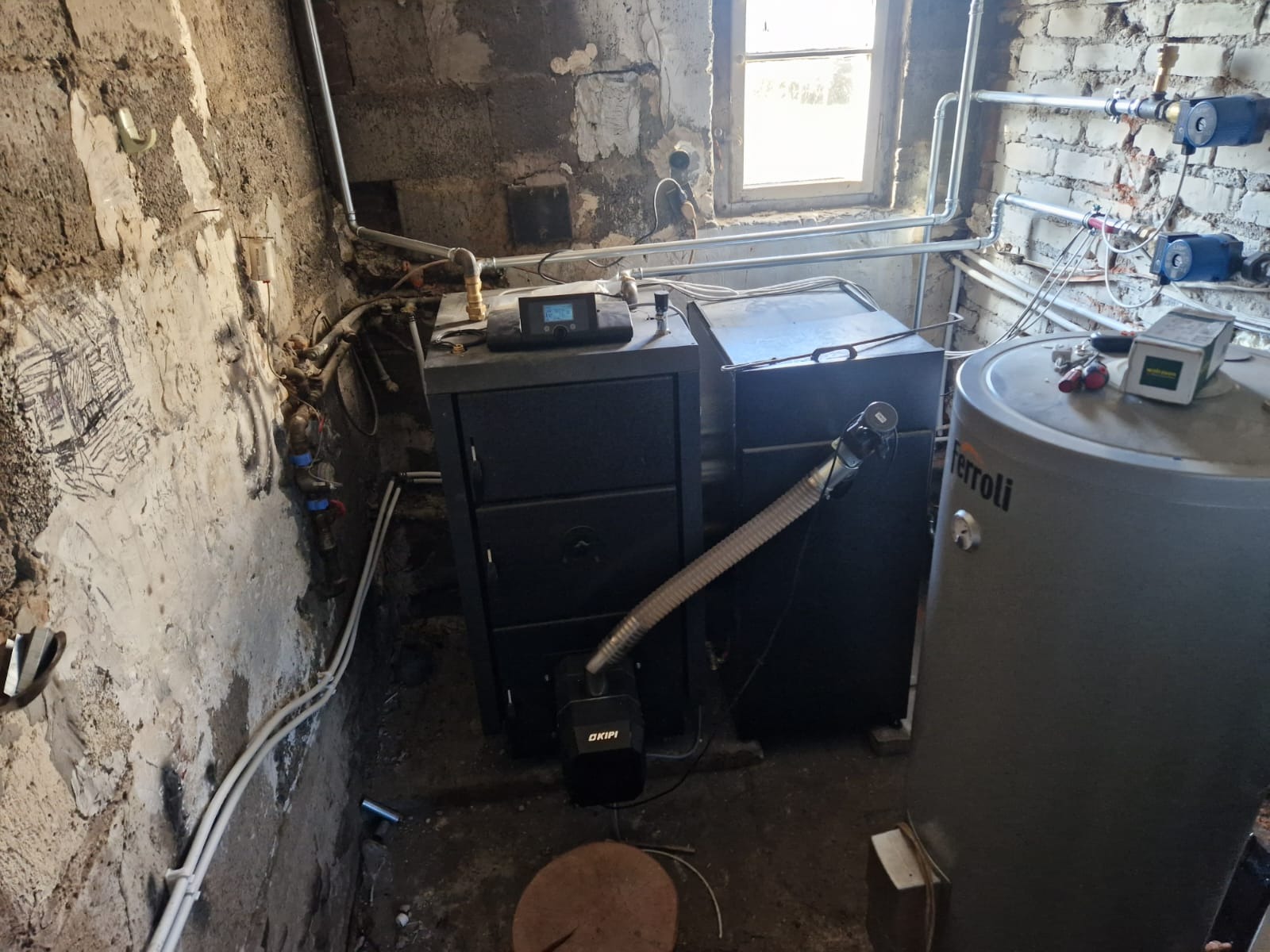 Czarny piec CO z podajnikiem i szary zasobnik ciepłej wody użytkowej Ferroli w surowym pomieszczeniu z instalacjami. Widoczne rury i okno.