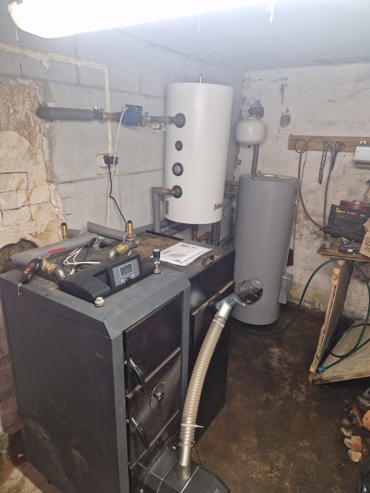 Kotłownia z piecem na paliwo stałe, zbiornikiem buforowym i instalacją CO w piwnicy z surowymi ścianami. Widoczne rury i zawory.