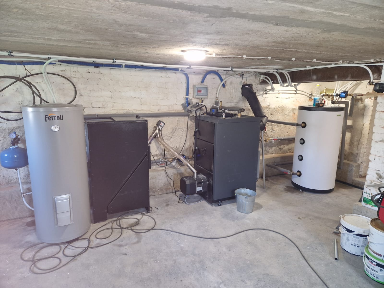 Kompletna instalacja CO w piwnicy z kotłem na paliwo stałe, zbiornikiem akumulacyjnym i zasobnikiem CWU marki Ferroli. Widoczne rury, przewody i osprzęt.