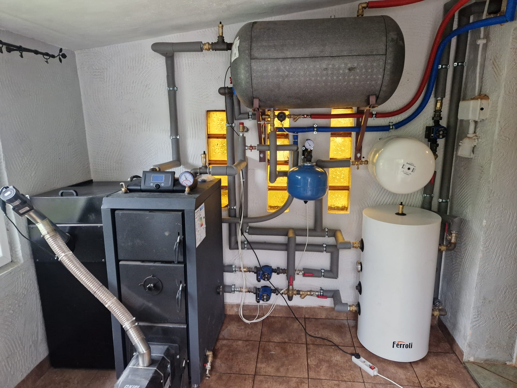 Kompleksowa kotłownia: piec na paliwo stałe z podajnikiem, zbiornik buforowy, zasobnik CWU Ferroli, naczynia przeponowe, rury w izolacji, armatura. Całość w pomieszczeniu z oknami z luksferów.
