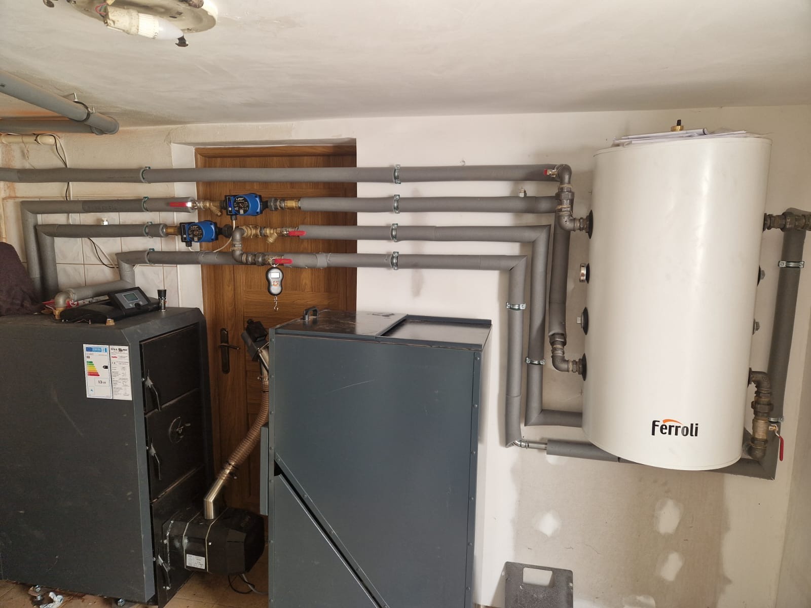 Kotłownia z piecem na paliwo stałe, zasobnikiem ciepłej wody Ferroli i izolowanymi rurami. Widoczne pompy obiegowe i zawory. Ujęcie w pomieszczeniu z drewnianymi drzwiami.