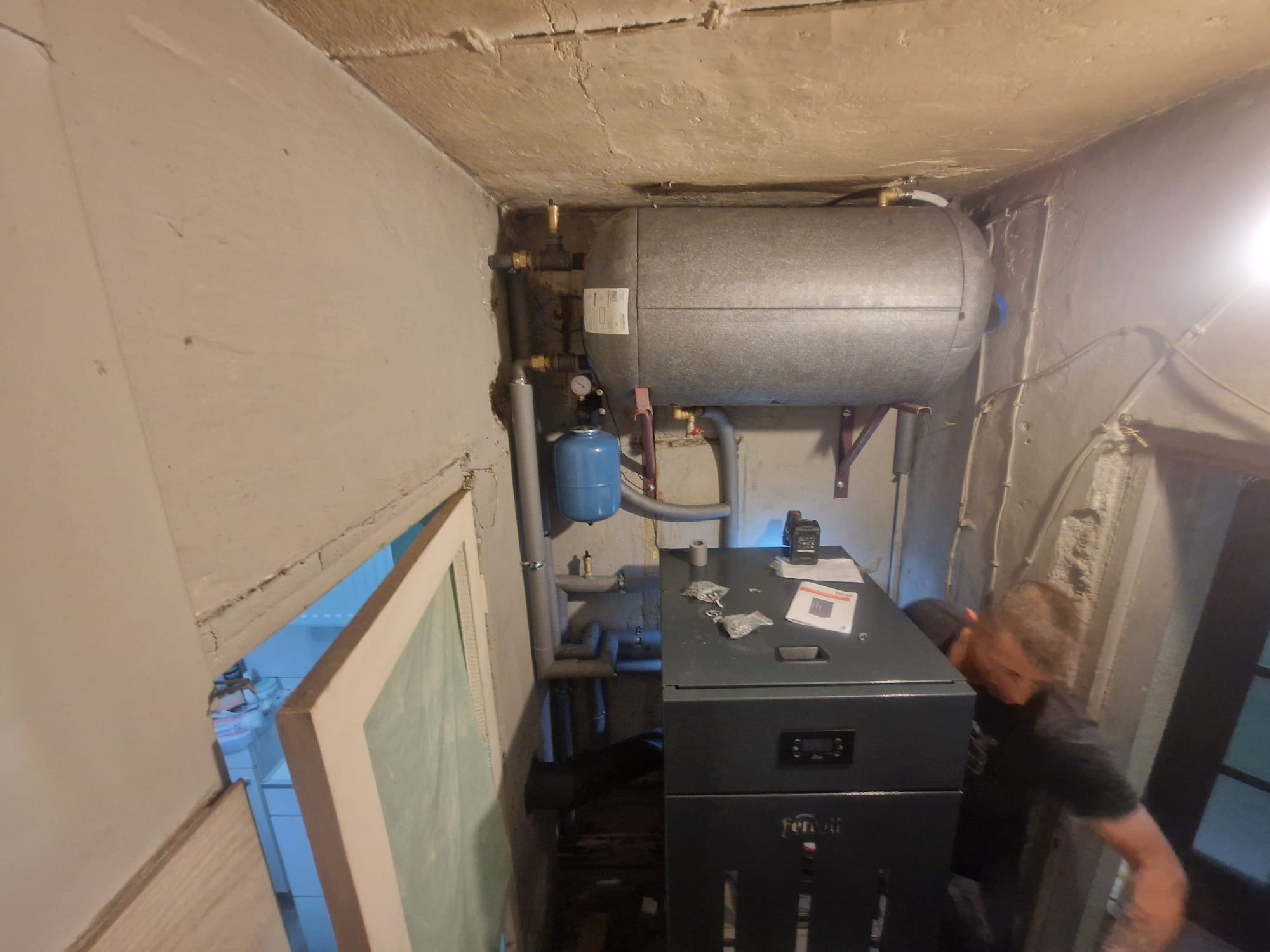 Szary piec gazowy Ferroli z podłączonym zasobnikiem wody w kotłowni. Widoczne rury, manometr i osoba podczas instalacji.