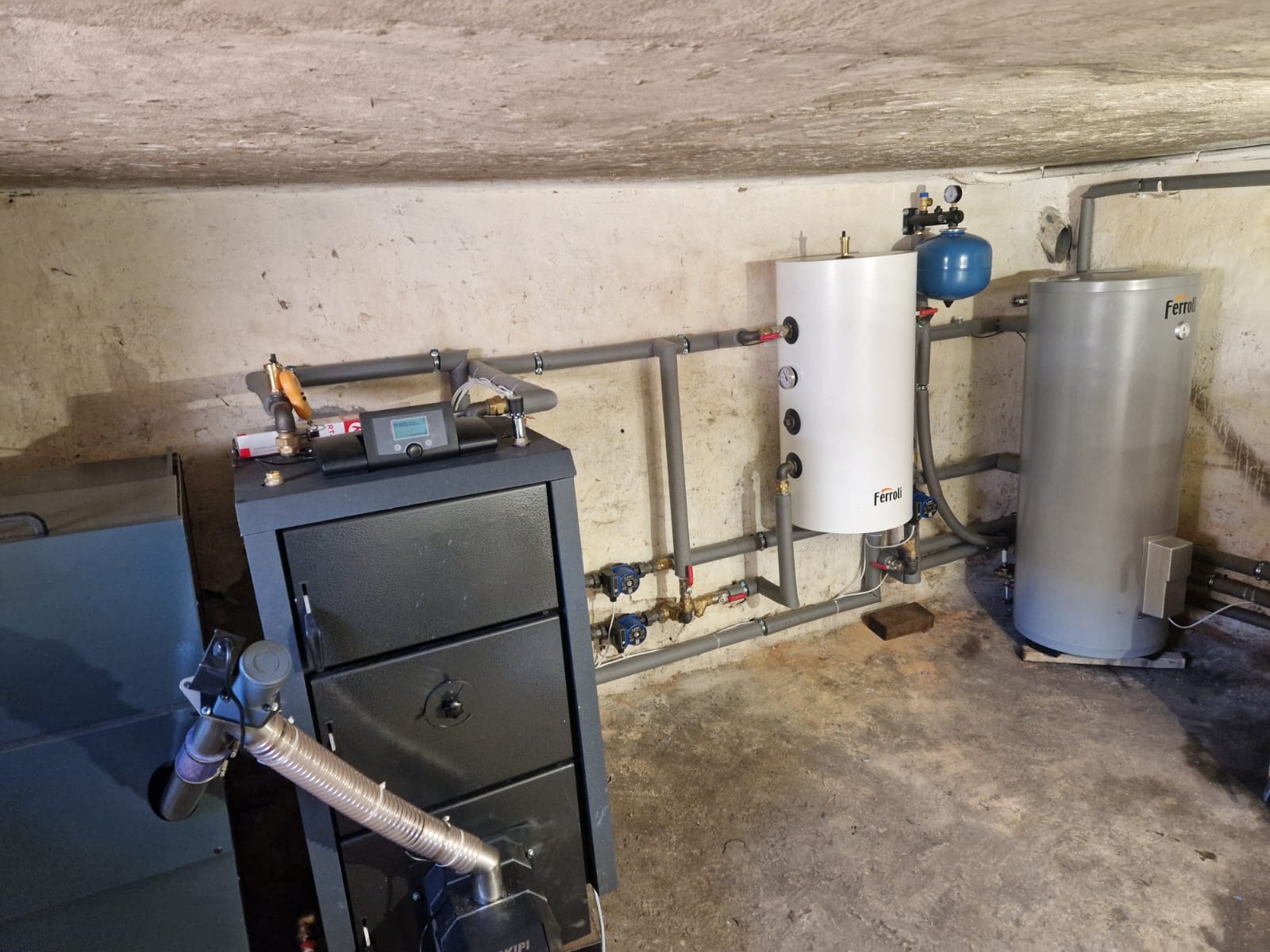 Kompletna instalacja grzewcza z piecem na paliwo stałe, zbiornikami Ferroli i szarymi rurami w piwnicy. Widoczne liczne zawory i manometry.