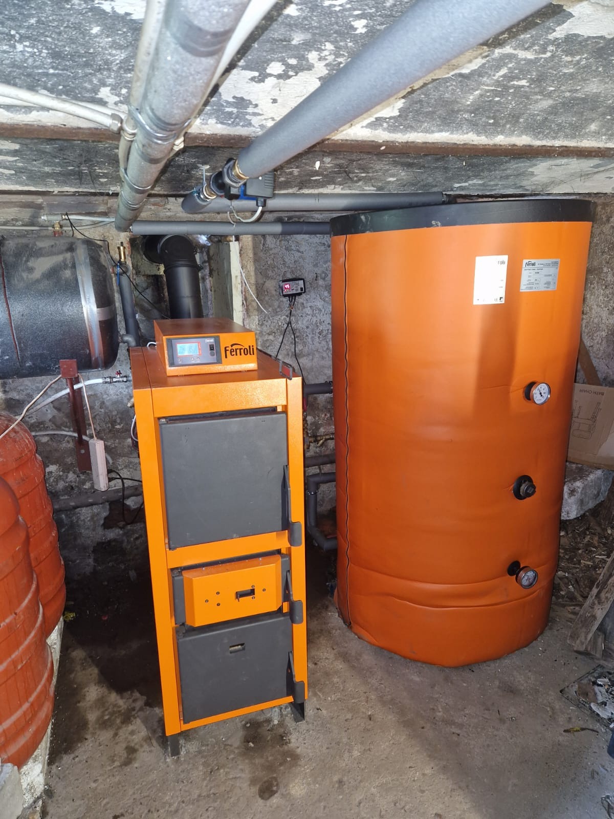 Pomarańczowy piec Ferolli z zbiornikiem buforowym w kotłowni. Widoczne rury instalacji CO. Surowy betonowy strop. Manometry na zbiorniku.