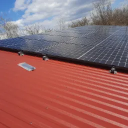 Panele słoneczne zamontowane na czerwonym dachu z blachy trapezowej, widoczne uchwyty montażowe i opakowanie po elementach instalacji.