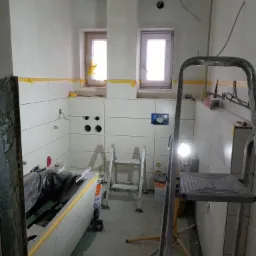 Łazienka w trakcie remontu: białe płytki na ścianach, przygotowane otwory pod instalacje, częściowo wykończony sufit z widocznymi łączeniami płyt, drabina malarska i narzędzia budowlane.