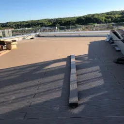 Rozległy taras na dachu z szarych desek, częściowo zacieniony, z metalowym profilem leżącym na powierzchni, szklane balustrady na krawędzi, widok na zalesione wzgórza w tle.