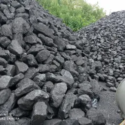 Dwa kopce węgla kamiennego o różnej granulacji, z fragmentem maszyny budowlanej widocznym w prawym dolnym rogu i zielenią w tle.