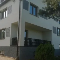 Dwupiętrowy dom z jasną elewacją, szarym kamiennym cokołem i drewnianymi panelami dekoracyjnymi wokół okien, widoczny z perspektywy ogrodu.