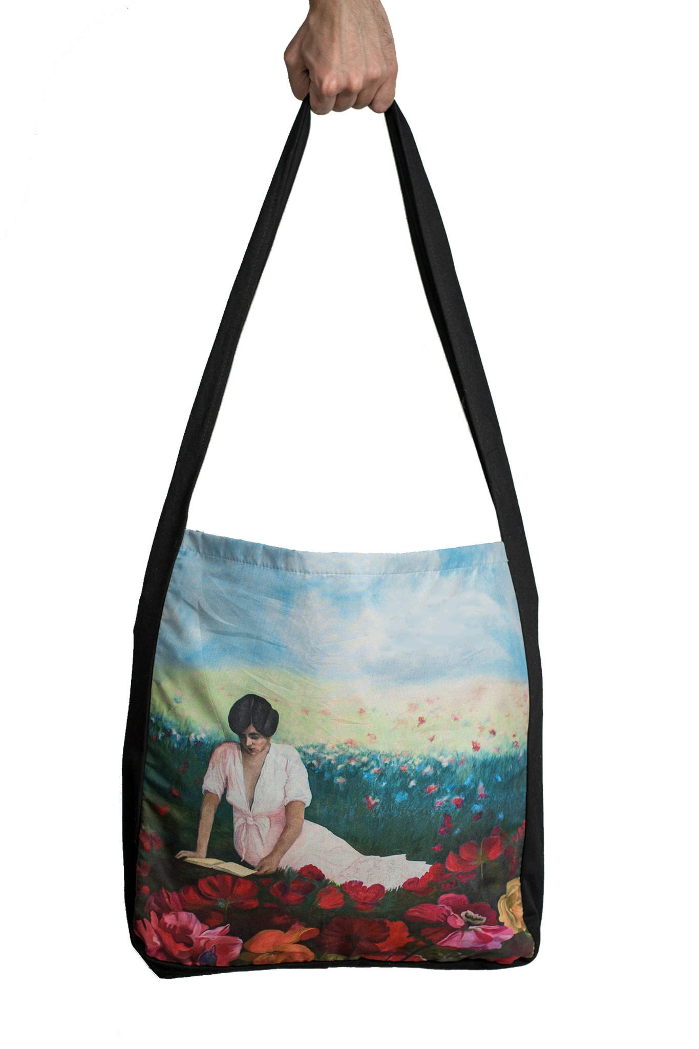 Torba na ramię z tkaniny z nadrukiem przedstawiającym kobietę czytającą książkę na łące kwiatów, trzymana przez rękę na białym tle.