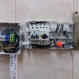 Otwarta skrzynka elektryczna z listwą zaciskową, obok skrzynka z wyłącznikami Eaton i Legrand oraz czerwone gniazdo siłowe Socomec na kafelkowanej ścianie.