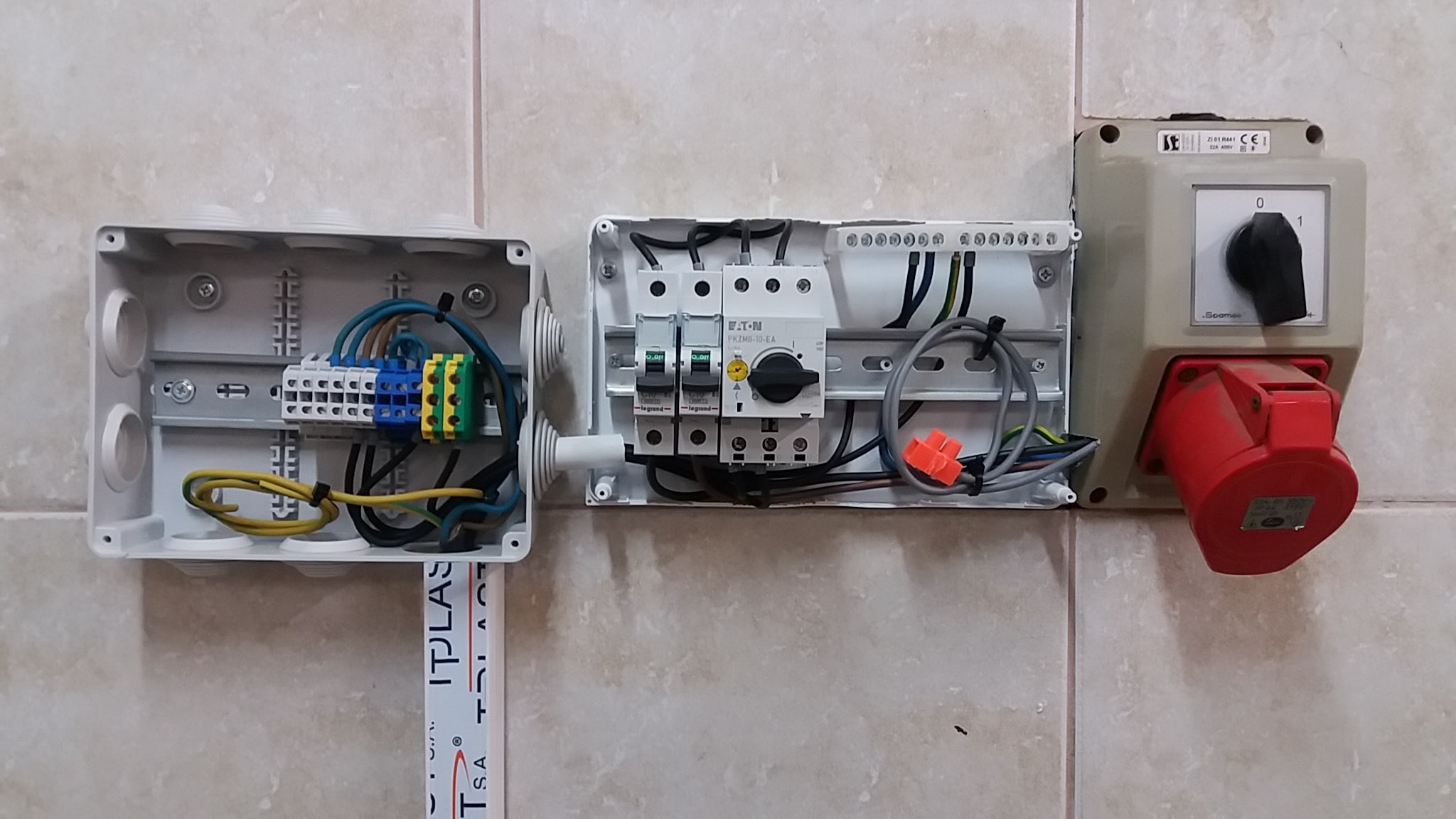 Otwarta skrzynka elektryczna z listwą zaciskową, obok skrzynka z wyłącznikami Eaton i Legrand oraz czerwone gniazdo siłowe Socomec na kafelkowanej ścianie.