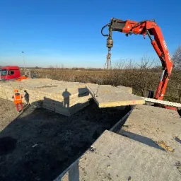 Czerwony dźwig Fassi podnosi prefabrykat betonowy na tle błękitnego nieba. W tle widoczny pracownik w pomarańczowym ubraniu i ciężarówki.
