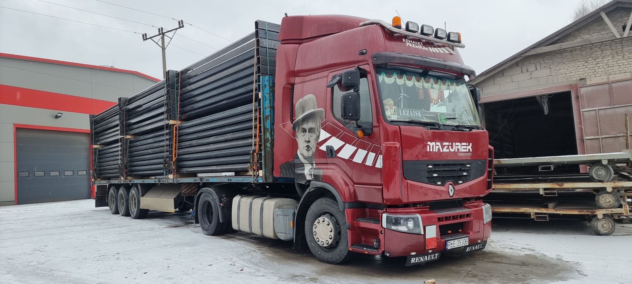 Czerwony ciągnik siodłowy z naczepą, przewożący ładunek stalowych profili. Na kabinie widoczny portret w kapeluszu. Zima, śnieg na ziemi.