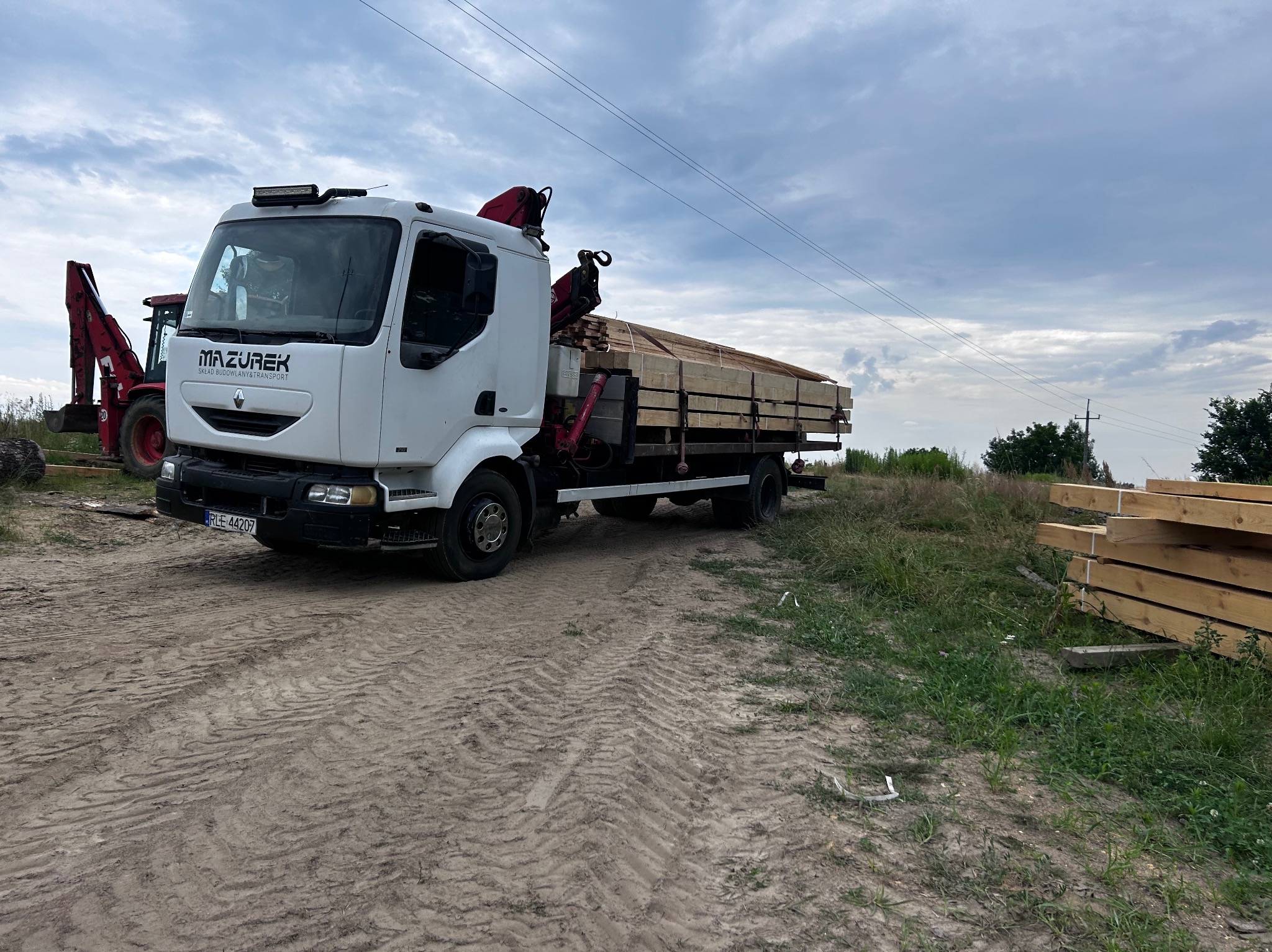 Ciężarówka Mazurek z dźwigiem i ładunkiem tarcicy na placu budowy, w tle czerwona ładowarka i ułożone drewno. Ślady opon na ziemi.