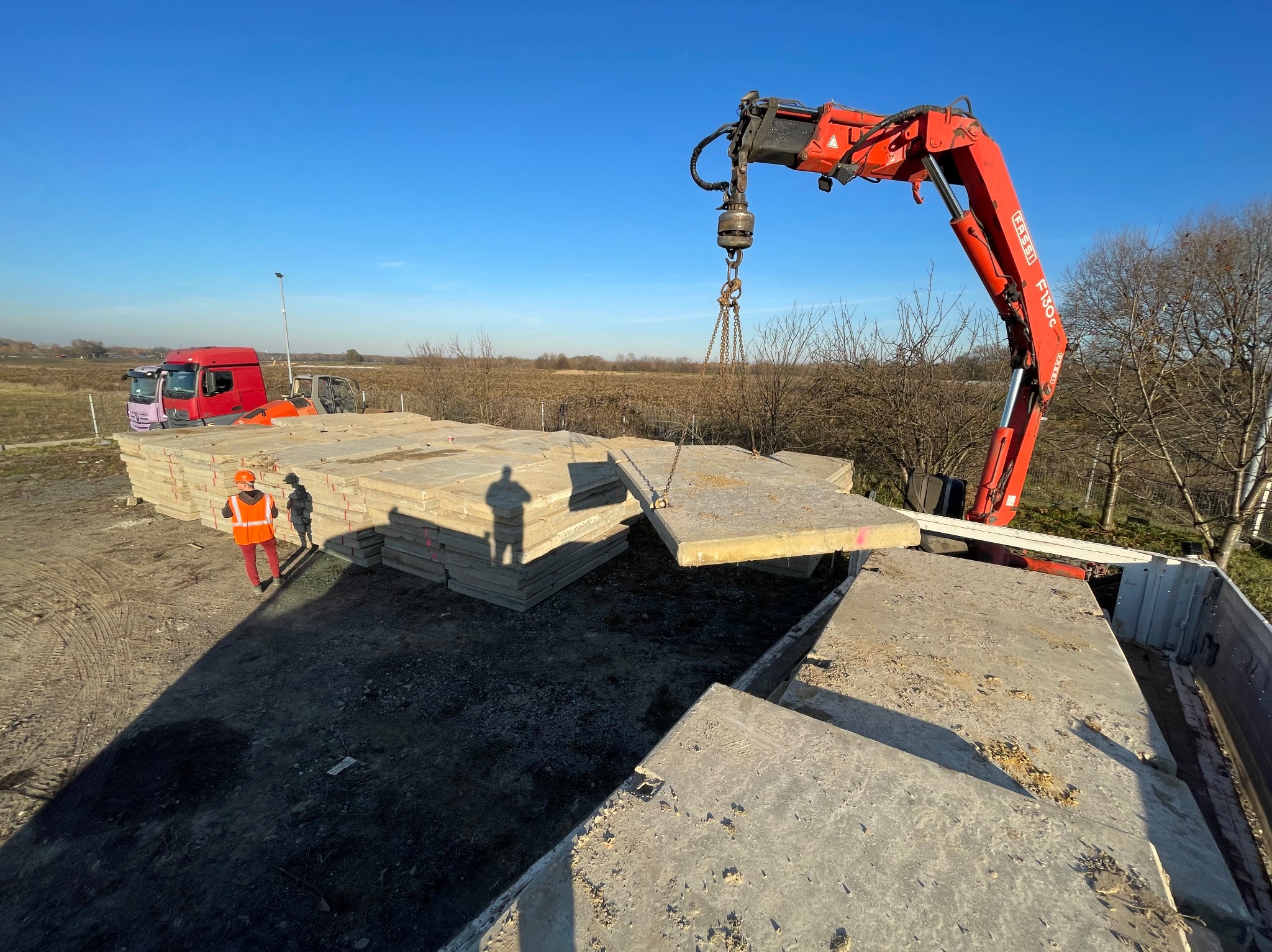 Czerwony dźwig Fassi podnosi prefabrykat betonowy na tle błękitnego nieba. W tle widoczny pracownik w pomarańczowym ubraniu i ciężarówki.
