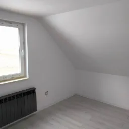 Poddasze po remoncie: białe ściany, jasne panele podłogowe imitujące drewno, czarny grzejnik pod oknem, widoczne gniazdko elektryczne na ścianie.
