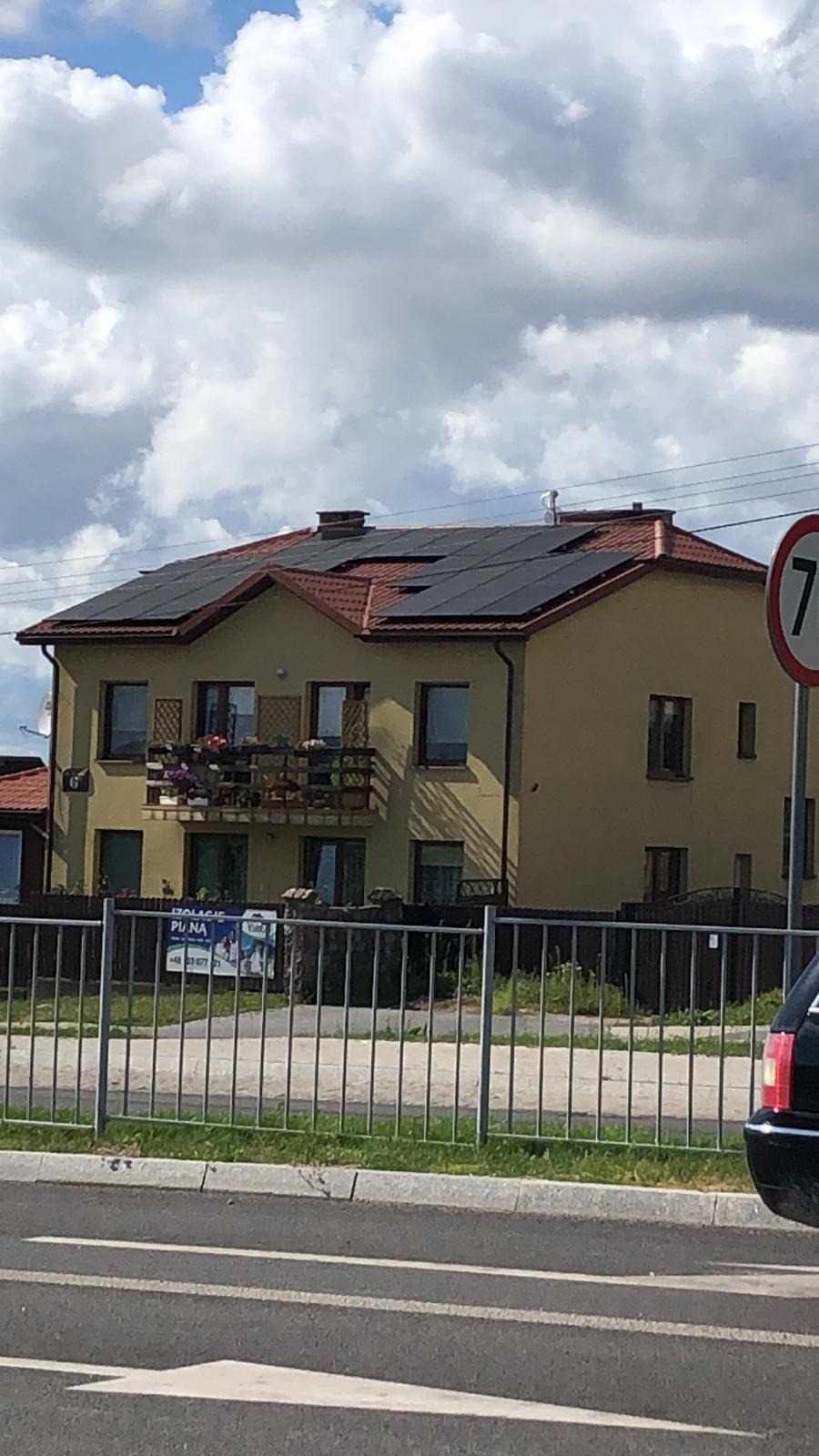 Dom jednorodzinny w Jabłonnie z zainstalowanymi panelami fotowoltaicznymi na dachu z czerwonej dachówki, widok z ulicy przez metalowe ogrodzenie.