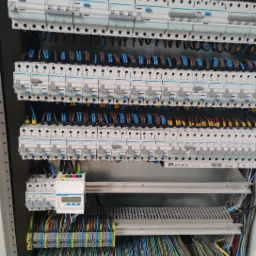 Rozdzielnia elektryczna z licznymi bezpiecznikami i okablowaniem w kolorach niebieskim, brązowym i żółtym, widoczny licznik energii.