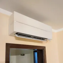 Biała klimatyzacja ścienna z otwartą klapą nawiewu, zamontowana nad ciemnobrązowymi drzwiami w beżowym pomieszczeniu.