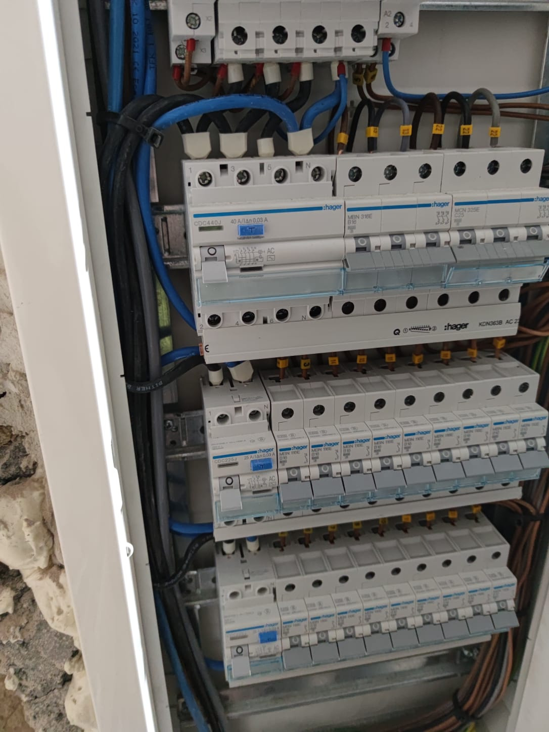 Rozdzielnica elektryczna Hager z zabezpieczeniami różnicowoprądowymi i nadprądowymi, widoczne okablowanie w kolorach niebieskim, czarnym i brązowym, zamontowana na ścianie.