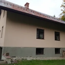 Dwukondygnacyjny dom z ocieploną elewacją w odcieniach beżu, z częściowo wykończonym cokołem w kolorze szarym, widoczne okna z ciemnymi ramami i metalowy dach z zabezpieczeniem przeciwśniegowym...