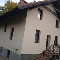 Dwukondygnacyjny dom z jasną elewacją, ciemnymi oknami i schodami wejściowymi z ozdobną balustradą, widoczne wykończenie fundamentów.