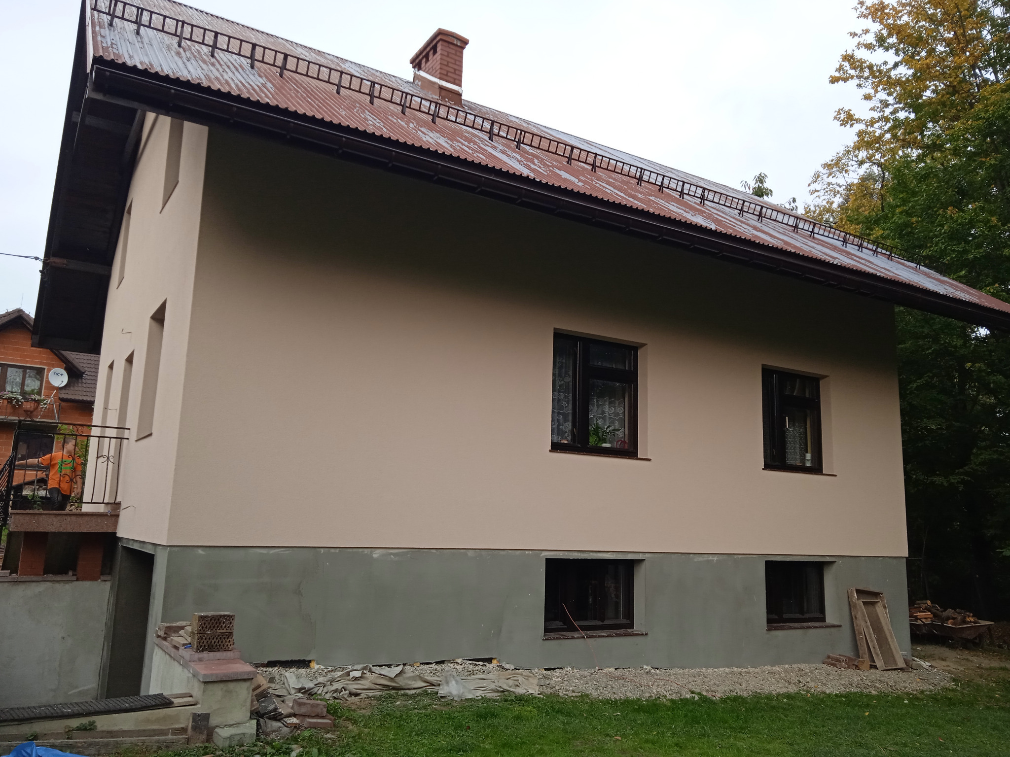 Dwukondygnacyjny dom z ocieploną elewacją w odcieniach beżu, z częściowo wykończonym cokołem w kolorze szarym, widoczne okna z ciemnymi ramami i metalowy dach z zabezpieczeniem przeciwśniegowym...