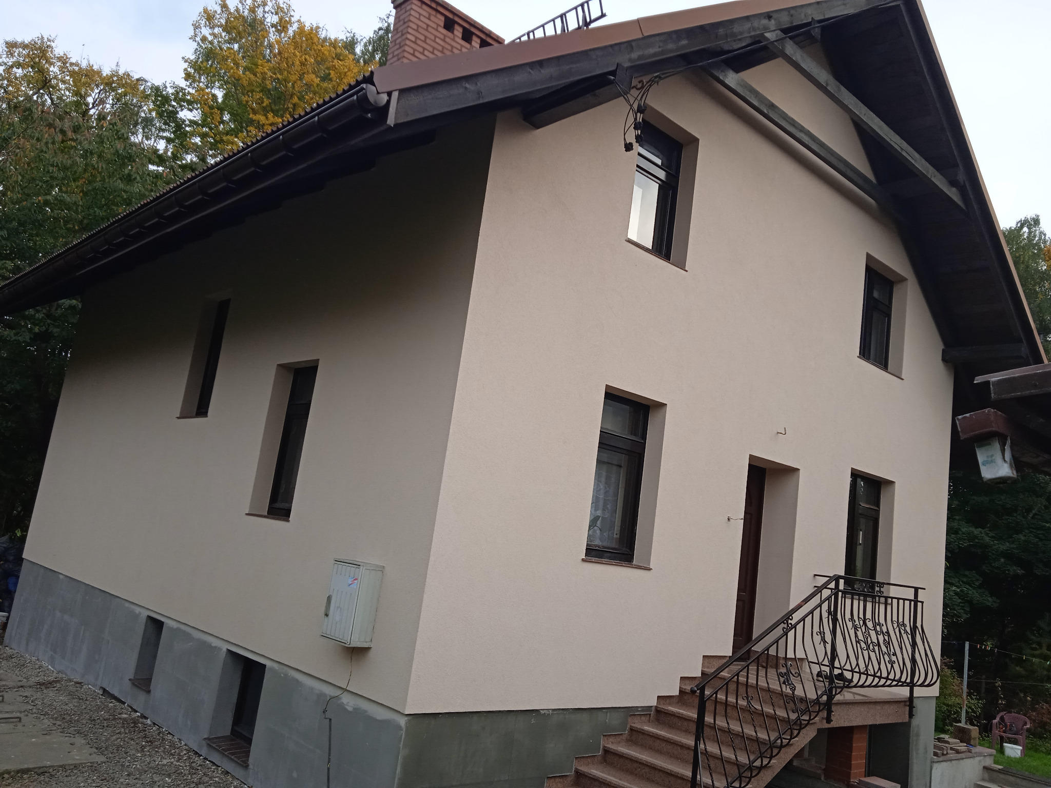 Dwukondygnacyjny dom z jasną elewacją, ciemnymi oknami i schodami wejściowymi z ozdobną balustradą, widoczne wykończenie fundamentów.