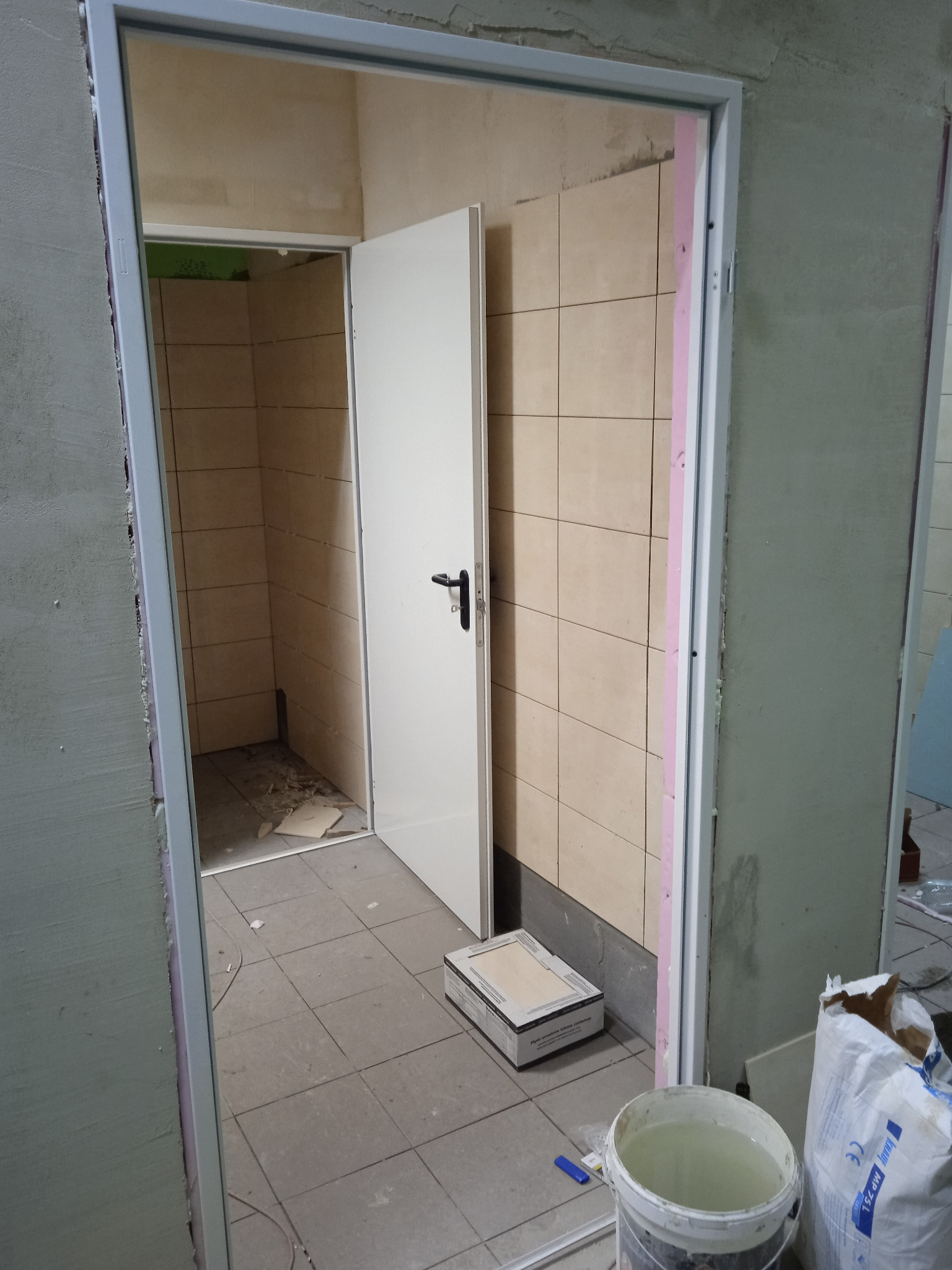 Łazienka w trakcie remontu: widoczne płytki na ścianach, białe drzwi z czarną klamką, wiadro z wodą, paczka płytek i worek z zaprawą na podłodze.