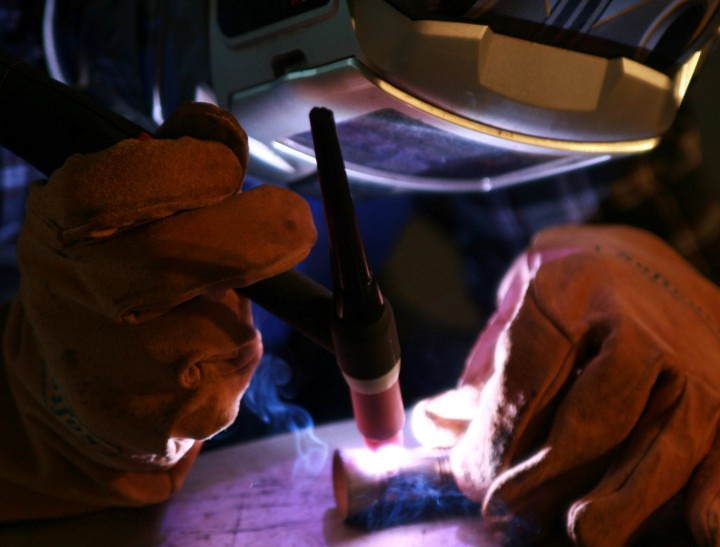 Spawanie rury miedzianej metodą TIG, widoczne iskry i dym, ochronna maska spawalnicza i rękawice robocze.