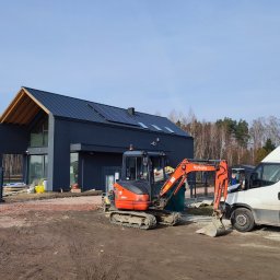 WM Your Home Waldemar Majek - Nowoczesny dom z panelami słonecznymi w Piasecznie, widoczny z placu budowy z mini koparką Kubota i samochodem dostawczym. Dom w stylu minimalistycznym.