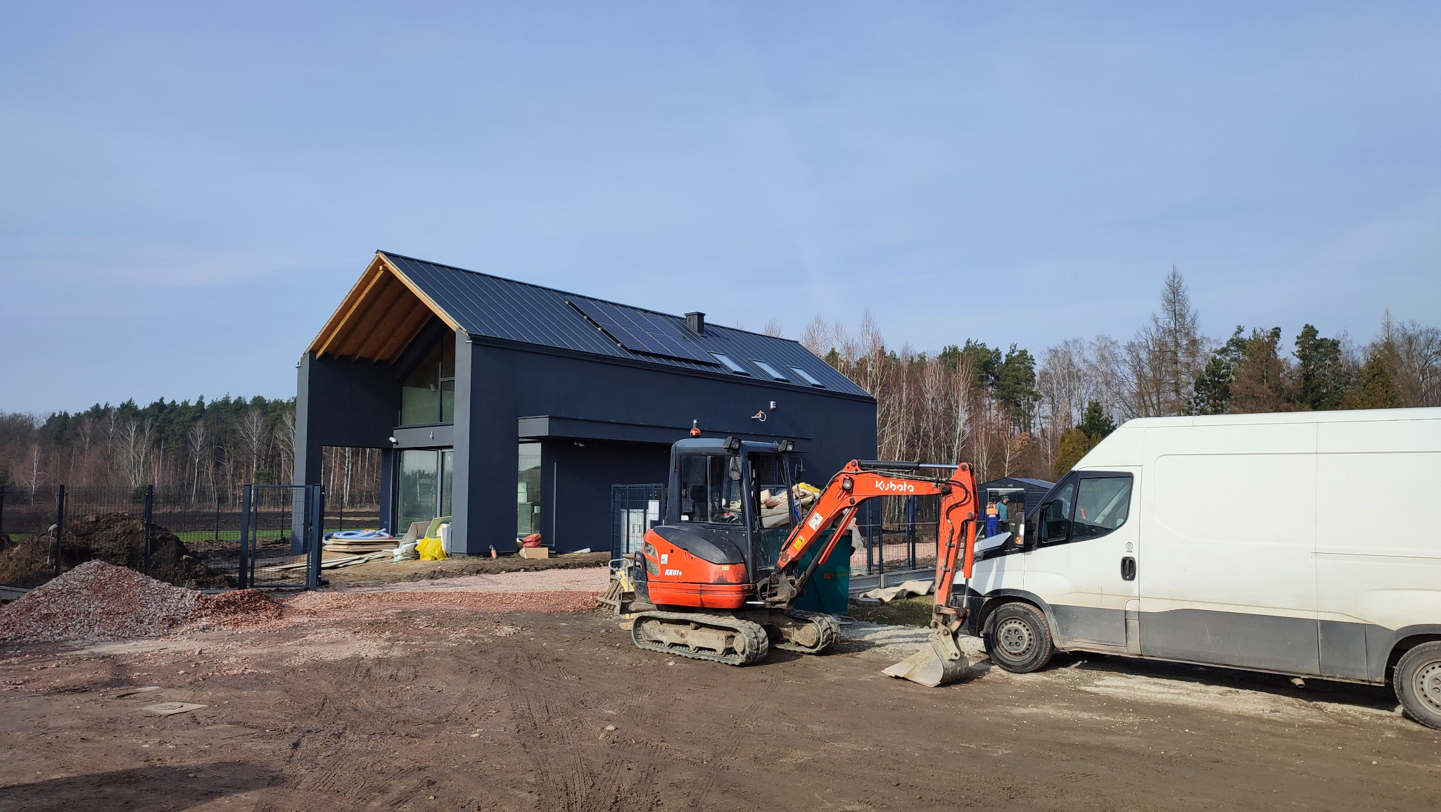 Nowoczesny dom z panelami słonecznymi w Piasecznie, widoczny z placu budowy z mini koparką Kubota i samochodem dostawczym. Dom w stylu minimalistycznym.