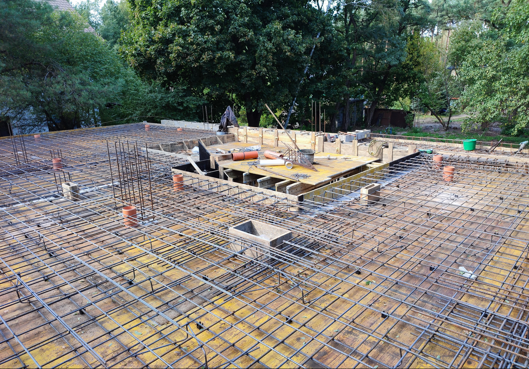 Zbrojenie stropu budynku jednorodzinnego z widocznymi elementami szalunków i instalacji, przygotowane do zalania betonem. Widok z góry na tle zieleni.